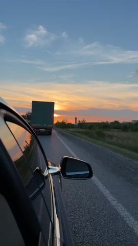 A car is traveling on a highway during sunset. The sun is low on the horizon, casting an orange and yellow glow across the sky, with scattered clouds. A large green truck is visible ahead on the same road, also traveling in the same direction. The side mirror of the car reflects the sunset. The environment appears to be rural, with fields and trees visible along the side of the road. The perspective is from inside the car, looking out the passenger side window.