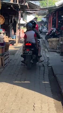 The video shows two people riding a motorcycle down a narrow alleyway, possibly a marketplace. The person in front is wearing a red shirt and a helmet, while the person behind is wearing a white shirt with red sleeves and a helmet. They are riding away from the camera, towards a section of the alley with more stalls and parked motorcycles. The alleyway is paved with interlocking bricks and lined with stalls on both sides selling various goods, some of which are woven baskets. The lighting suggests it is daytime, with sunlight filtering through an overhead covering.