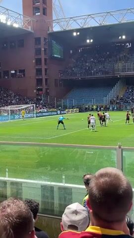 The video shows a soccer match from the stands. In the foreground, heads of spectators are visible, some wearing hats. Through a clear barrier, the green soccer field is in view, with players wearing white and red/black uniforms engaged in play. A referee in black is positioned on the field. The goal is visible to the left, and advertisements surround the field. Spectators fill the stands of the stadium. A large screen displays the game. The weather is sunny. At one point, the ball goes into the net, eliciting cheers from the crowd. A man in a green safety vest walks by the barrier.