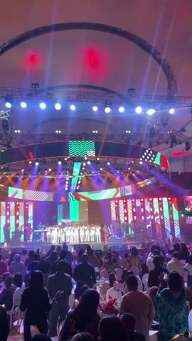 An indoor event is occurring in a large, spacious hall. The room is filled with numerous seated guests, many of whom are standing and observing a performance on a stage. The stage is elaborate, featuring colorful light displays, a large screen with geometric patterns, and a choir or group of performers dressed in white outfits. A band is playing instruments. The stage is decorated with colorful geometric shapes. Several stage lights illuminate the stage. The overhead structure includes a large circular element that has lights on it. Many attendees are holding up their phones, presumably to record or take photos of the event. The atmosphere is vibrant and festive.