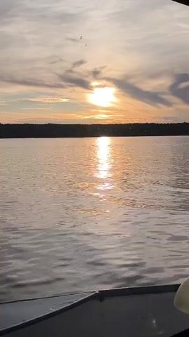 A serene sunset is viewed from a boat on a body of water. The sun is partially obscured by clouds, casting a warm glow across the sky and reflecting on the water. The horizon is lined with trees. A portion of the boat's railing is visible in the foreground.