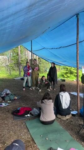 This media file captures a group of people gathered outdoors under a large, draped blue tarp, likely for an activity or class in a natural setting. The ground is covered in dirt and pine needles, with trees visible in the background, suggesting a forest location. Several individuals are standing under the tarp, appearing to be participants or instructors. One person in a tan uniform holds a tablet, seemingly leading the group. Another individual in a green jumpsuit is striking a pose, with two others in darker clothing also participating in some form of movement or exercise. In the foreground, several people are seated on the ground, some on yoga mats, facing the standing group. This arrangement suggests a workshop, a class, or an informal gathering focused on instruction or demonstration.

Objects visible include yoga mats, backpacks, and what appear to be papers or notebooks scattered on the ground. The lighting suggests it is daytime, with diffused light filtering through the tarp and trees, indicating a possibly overcast or shaded environment. The overall atmosphere seems relaxed and engaged, with participants focused on the activities. The mention of "Soanlakhti, Russia" provides geographical context for the scene.