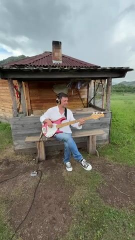 A man wearing headphones, a white shirt, blue jeans, and black and white sneakers sits on a wooden bench in front of a rustic wooden structure. He is playing a pink bass guitar. A small black electronic device and a black cable are on the ground to his left. The structure has a wooden roof and a brick chimney. The background is a grassy field with hills and a cloudy sky.