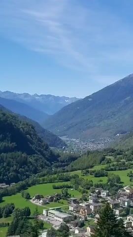 The video captures a sweeping panoramic view of a mountainous valley on a clear, sunny day. Lush green forests cover the slopes of the mountains, which recede into the distance, becoming progressively hazy and blue. Below, a valley floor is dotted with small towns and villages, with agricultural fields of vibrant green interspersed throughout. The scene is tranquil and serene, showcasing the natural beauty of a mountainous landscape.