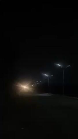 The video shows a dark scene, illuminated by streetlights and the headlights of an approaching car. The streetlights are spaced along the side of the road. The road itself is dark. The car's headlights are bright, and the vehicle appears to be driving towards the camera.