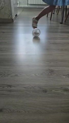 A clear, round ball with a small creature inside rolls across a wooden floor. The ball reflects light, creating a bright spotlight on the floor in front of it. The floor has a grey, wood-grain pattern. In the background, the legs of chairs are visible, along with a radiator and what appears to be a kitchen counter. The scene is well-lit, suggesting it's daytime. The overall impression is one of a domestic setting, possibly a kitchen or dining area in a home. The creature inside the ball is too small and indistinct to identify specifically, but it appears to be moving within its transparent enclosure.
