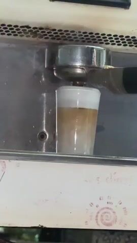 A close-up video shows a coffee machine dispensing a drink into a clear glass cup. The drink is layered, with white foam on top, a darker brown liquid below it, and a lighter brown liquid at the bottom. The coffee machine's portafilter is attached, and the hot liquid is flowing through it into the cup. The machine appears to be metal, with a grid-like vent at the top. There are some decorative elements on the white surface below the machine, which appear to be stamps of a spiral design. The lighting is overhead and appears to be artificial. The video is brief, lasting only a few seconds.