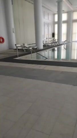 The video pans across an indoor swimming pool area. The pool is rectangular and appears to be a lap pool. There are lounge chairs on either side of the pool, and some exercise equipment, including stationary bikes, is visible to the right. Large windows with blinds cover the far wall, letting in diffused light. The floor is tiled with large, light-colored tiles. The overall atmosphere is clean and modern. There are no people visible in the video.