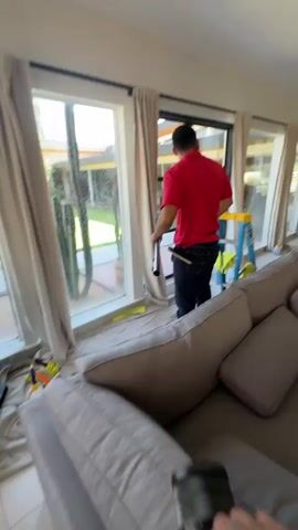 The video opens with a shaky, blurry shot of a hand holding a phone, with a man in a red shirt visible in the background near a window and curtains. The perspective shifts to a cleaner in a dark blue shirt, kneeling on the floor in front of a large window. He is applying some kind of film or tint to the glass. There is a bucket with cleaning supplies next to him, along with some tools and cloths spread on the floor, indicating ongoing window cleaning or application work. Outside the window, there is a swimming pool, a grassy yard, and a modern-looking house. The sun is shining, suggesting it is daytime. The hand holding the phone reappears briefly, and then the view focuses on the cleaner's work.