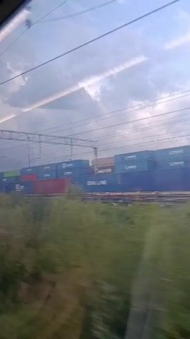 The video shows a side view from a moving vehicle, likely a train, looking out a window. The foreground is blurred with green foliage, indicating rapid movement. In the mid-ground, a long freight train is visible, composed of numerous stacked shipping containers. The containers are various colors, including blue, green, and red, with some bearing company logos or text such as "MAERSK" and "DRA LINE". Overhead, a complex network of power lines and support structures for an electric railway is apparent against a cloudy sky. The lighting suggests it is daytime, possibly overcast or nearing dusk, with streaks of light breaking through the clouds. There are no people directly visible in the frame.