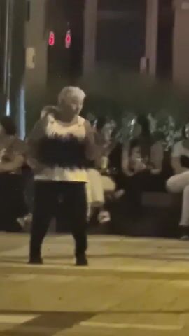 In this video, an elderly woman with short, white hair is dancing in the street at night. She is wearing a sleeveless tank top with a black and white tie-dye pattern and black pants. She is in the middle of a crosswalk, with other people sitting on a bench behind her. The scene is dimly lit, suggesting it is nighttime. The atmosphere appears festive, with the woman's energetic dancing and the presence of an audience. The background shows a building with illuminated windows and what appear to be traffic lights, indicating an urban setting. The woman's movements are lively and expressive, suggesting she is enjoying herself. The presence of other people watching her implies a community gathering or performance. The overall impression is one of a spontaneous street celebration.
