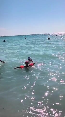 A video shows two people swimming in the ocean on a sunny day. The water is clear and blue, with sunlight glinting off the surface. There are other people in the distance, also swimming and enjoying the water. The person in the foreground is wearing a red flotation device, while the person behind them is wearing a blue cap and a red flotation device. Both individuals appear to be relaxed and happy as they swim. The scene suggests a typical day at the beach, with people enjoying the water and sunshine.