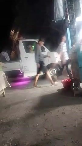 At nighttime, a white pickup truck is parked on a dirt road. The truck has underglow lighting that emits a pinkish purple hue. Several people are near and inside the truck. In the bed of the truck, two individuals are visible, one in red clothing and another in a red and yellow outfit. On the driver's side of the truck, a person is seated in the driver's seat. Outside the truck, on the driver's side, two people are walking. One person is wearing a striped shirt and shorts, and the other person is wearing an orange vest and jeans. To the left of the truck, a table with items, including a large water jug and a box, is set up. To the right of the truck, a stall-like structure with items displayed on it is present, along with a red bucket and a small stool. The ground is covered with dirt and scattered leaves.
