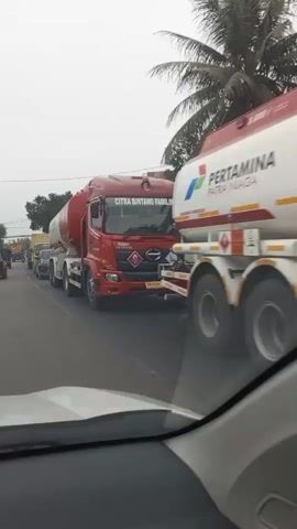 The video shows a line of vehicles on a road. In the foreground, the camera appears to be inside a vehicle looking out through the windshield. The first truck visible is a red tanker truck with "CITRA BINTANG FAMILINDO" written on the side. Behind it, there is another tanker truck with "PERTAMINA PARA NIAGA" written on it. Following these trucks are several other vehicles, including cars and smaller trucks, all moving slowly in what appears to be a traffic jam. The environment seems to be outdoors with trees and buildings visible in the background. The sky is overcast. There are no clearly discernible people in the video.
