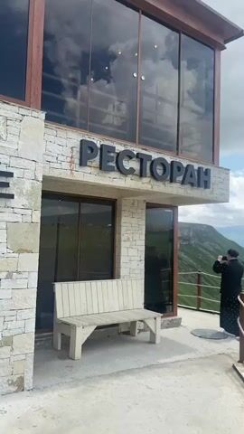 A woman stands on a concrete patio, facing away from the camera, holding a smartphone to her face as if taking a picture. She is wearing a dark headscarf and a dark, patterned dress. To her left is a white wooden bench. Behind the bench is a building with large windows and the word "РЕСТОРАН" in large black letters. The building appears to be made of light-colored stone. To the right of the building, a railing separates the patio from a steep, grassy cliff. The cliff drops into a deep canyon with a river at the bottom. In the distance, there are rolling green hills and mountains under a cloudy sky.