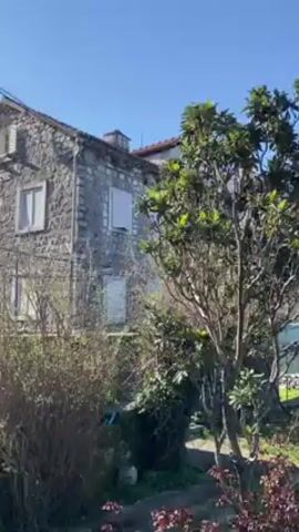 The camera pans left and upward to show a three-story stone house with white-framed windows and shutters. A small balcony with a black metal railing is attached to the front of the house. There is a clear blue sky in the background. Trees and flowering bushes are in the foreground. Behind the house, the top of a chimney is visible, and mountains are in the distance.
