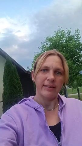 A woman with blonde hair and a purple hoodie is filmed from the chest up, looking directly at the camera. She appears to be outside, with greenery and the sky visible behind her. The lighting suggests it may be dusk or dawn.