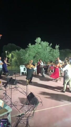 A group of people are dancing on a patio at night. In the foreground, a couple in formal attire is dancing the tango. The woman is wearing a black strapless dress and the man is wearing a black suit. Behind them, another couple is dancing. The woman is wearing a burgundy dress and the man is wearing a white shirt and dark pants. Several other people are visible in the background, some watching the dancers and others standing around tables. The environment appears to be an outdoor venue with trees and lighting.