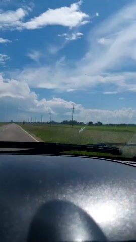 The video shows a view from inside a car looking out the windshield. A paved road stretches into the distance, bordered by green fields and utility poles. The sky is blue with scattered white clouds, and the sun is casting bright reflections on the car's dashboard. The wiper blades are visible in the foreground. There are no people or other vehicles visible.