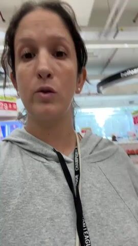 A woman with brown hair pulled back and wearing a light gray hooded sweatshirt is standing in a supermarket aisle. She is pointing to the right with her right hand. The aisle is stocked with shelves of products. Other shoppers can be seen in the background. The woman is smiling.