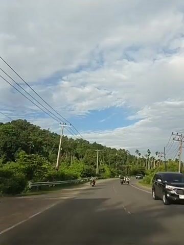 The video shows a paved road in a tropical or subtropical region. The road curves to the right, with lush green vegetation and trees on both sides. In the distance, there are hills covered in dense foliage. Several utility poles with power lines are visible along the roadside. There are a few motorcycles on the road, suggesting light traffic. The sky is partly cloudy with a mix of blue sky and white clouds. The overall atmosphere is serene and natural.