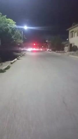 The video shows a street at night. There is a streetlight on the left side of the road, and a building on the right. A vehicle with bright headlights is approaching the camera, causing glare. The camera appears to be mounted on a bicycle, as a wheel is visible in the foreground.