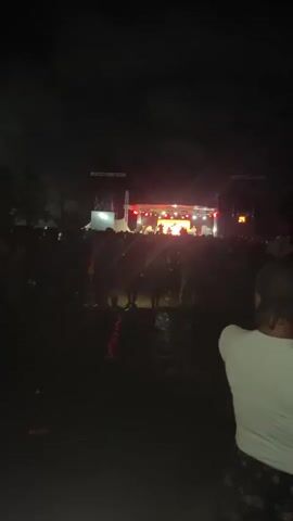 At night, an audience watches a performance on a stage. The stage is decorated with red and white lights, and it features a screen with a round logo. Several performers, some standing and some seated, are illuminated on stage. The audience is mostly silhouetted against the light of the stage, with a few individuals more visible in the foreground. A man with a white shirt is seen on the right corner of the frame, facing the stage. Beams of light shoot up into the dark sky.