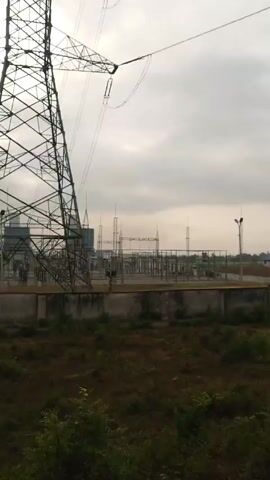 The video shows an electrical power station under a cloudy sky. In the foreground, there is a tall power transmission tower made of interconnected metal beams. Behind the tower, and separated by a concrete wall, lies the power station itself, featuring multiple structures with electrical components and wires. Further in the background, there are three cooling towers with red and white striped tops. The ground in front of the wall is covered in overgrown grass and shrubs. The scene is static with no discernible activity.
