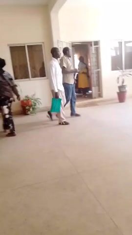 The video shows the entrance to the Department of Social Science Education at a building in Nigeria. Two men walk into the building at the beginning of the video. Later, two women enter the same entrance. The room is brightly lit with windows and plants in pots around the entrance. There is a table with a green bucket on it just outside the door. The video appears to be recorded on a mobile device.