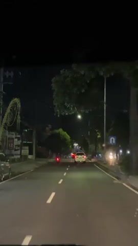 The video captures a night-time drive from the perspective of a vehicle's dashboard, looking forward onto a road in Kelurahan Lateng, Indonesia. The road is illuminated by several streetlights, with the sky appearing dark. The asphalt road features white dashed and solid lane markings, clearly visible under the artificial light.

Trees line both sides of the road, their dark silhouettes and some green foliage noticeable. On the left, vague outlines of buildings or structures are seen in the darkness. Ahead, a white car is driving in the same direction, its red taillights glowing. To the left of the white car, a red motorcycle also proceeds forward. Further down the road, additional vehicle lights are visible, indicating other traffic.

On the right side of the road, a blue rectangular sign with a white U-turn arrow is mounted on a pole. The scene conveys a quiet drive through an urban or suburban area at night, with no specific weather events like rain or fog apparent.