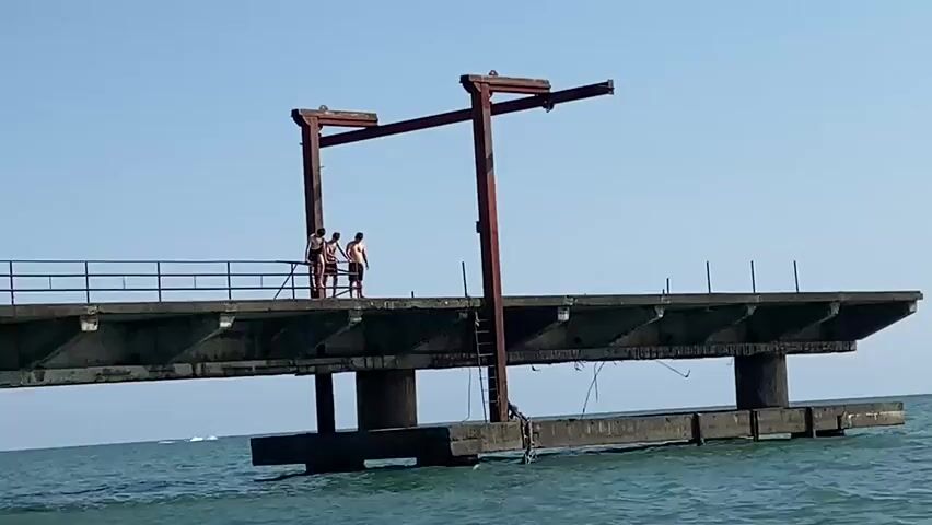Three young men stand on a concrete pier over the ocean. They are wearing shorts and t-shirts. One of them appears to be holding a fishing rod. The pier has metal railings and a rusted metal structure above it, resembling a gantry crane. The ocean is a calm blue, and the sky is clear.