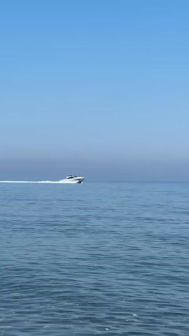A white speedboat is cruising across a vast expanse of blue ocean. The boat is angled slightly to the left of the frame, creating a white wake behind it that stretches across the water. The sky above is a clear, bright blue, transitioning to a slightly hazy, lighter blue near the horizon. The water is a deep blue with gentle ripples on the surface. The video is shot from a low angle, possibly from another boat or the shore. The overall scene is one of peaceful movement and open water.