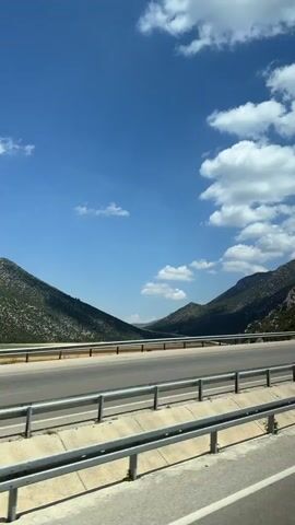 The video shows a scenic view of a road passing through a valley. The sky is clear with some fluffy white clouds. On the left side of the road, there are mountains covered with green trees. On the right side, there are also mountains. Cars are driving on the road. One dark-colored SUV is in the left lane. Another car, a white van with a logo on the side, is in the right lane. There are guardrails along the side of the road. The road is paved and appears to be a highway. In the foreground, there are some trees and bushes. The overall impression is a beautiful, sunny day on a road trip through a mountainous landscape.