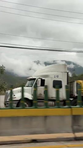 A dashcam view from a moving vehicle captures a cloudy day in a mountainous region. The video shows a truck with blue and yellow tarps, a green and yellow barrier, houses, and power lines. The vehicle is moving, indicated by the passing scenery.