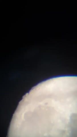 The media file is a close-up, slightly out-of-focus view of the Moon against a dark sky. The image is dominated by the Moon's surface, which shows craters and textured features. The curvature of the Moon is visible, with its illuminated portion taking up the majority of the frame. The edge of the Moon shows a slight rainbow-like chromatic aberration, suggesting the image was captured through a lens or telescope. The upper portion of the frame is black, indicating a night sky. There is no visible text in the media file. Given the location context of Târgu Neamț, Romania, this depicts an astronomical observation of the Moon at night. No people, specific objects other than the Moon, or distinct weather conditions are discernible. The activity is passive observation of a celestial body.