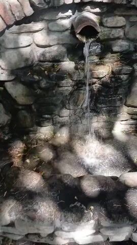 The video shows a close-up of a small man-made waterfall. Water flows from a ceramic pipe embedded in a stone wall covered with moss and some leaves. The water cascades down a rock formation into a pool below. The camera then pans slightly to the right, revealing a larger stone structure with a brick arch. In the background, there is a fan, a fire extinguisher, and a barbecue grill with some corn. The environment appears to be outdoors, possibly a garden or an outdoor cooking area. The lighting suggests it is daytime, with sunlight filtering through. There are no people visible in the video. The overall impression is of a rustic, functional outdoor space with a decorative water feature.