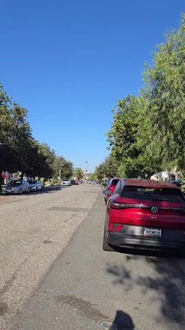 A first-person perspective video shows a residential street lined with trees and parked cars under a clear blue sky. The camera pans slightly to the right, revealing a red Volkswagen ID.4 parked on the right side of the street, followed by a line of other vehicles. The street surface appears to be asphalt with some oil stains. On the left side of the street, houses are visible behind a row of trees, with more parked cars. The video captures a typical suburban scene on a sunny day.