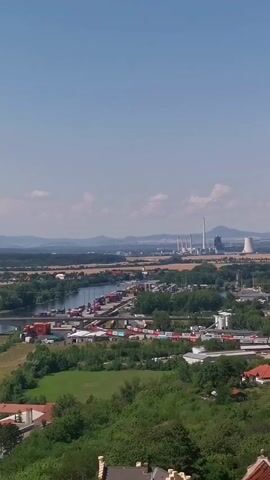 A sweeping aerial view captures a sunny landscape with a clear blue sky and scattered clouds. In the mid-ground, a wide river is bordered by a busy port with numerous shipping containers and trains. Beyond the port, a power plant with several tall smokestacks and a cooling tower stands prominently against the horizon. The surrounding area is a mix of green fields, lush trees, and scattered buildings, including some with red-tiled roofs, suggesting a rural or semi-urban environment. The foreground features the tops of buildings with terracotta roofs, indicating the vantage point is from a higher elevation. The overall impression is one of industrial activity coexisting with natural scenery under a bright, clear sky.