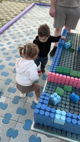 Two young children are playing with large plastic blocks on a tiled outdoor surface. A girl with pigtails, wearing a white jacket and brown pants, holds a red block. A boy in a black jacket and jeans stands next to her, leaning on a structure made of the blocks. An adult is partially visible in the background, wearing shorts and dark shoes. The tiled surface has a blue and white pattern. The blocks are various colors, including blue, pink, and green.