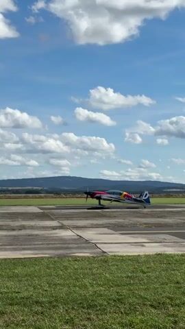 A small, colorful airplane, branded with "Red Bull" and a bull logo, sits stationary on a concrete runway. The plane is predominantly blue with red and pink accents on its wings and fuselage. It has a propeller at the front. In the distance, rolling green hills and a hazy blue sky with scattered white clouds form the backdrop. A second, much smaller aircraft is visible in the sky in the upper right quadrant of the frame. The foreground consists of a strip of green grass. The video appears to be taken from ground level, looking across the runway towards the airplane and the distant landscape.