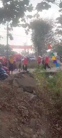 The video shows a street scene with a procession of motorcycles and people. There are flags with various colors, including red, white, blue, yellow, and green, displayed on poles. Some of the people are wearing red clothing, and others are wearing headscarves. There are trees and vegetation in the background, as well as buildings and power lines. The scene appears to be a festive event, possibly a parade or celebration. Some of the motorcycles are adorned with flags. A banner with Indonesian writing is visible in the distance, suggesting a connection to Indonesia. The road is paved, and there are some rocks and debris on the side of the road. The weather appears to be overcast or hazy.