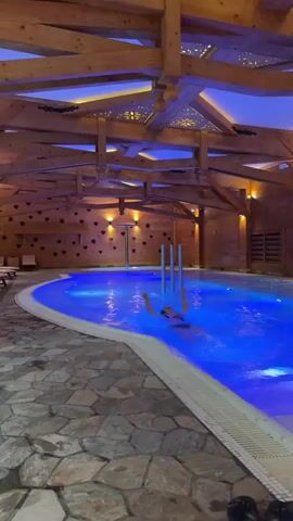 This video captures an indoor swimming pool area, likely within a spa or hotel, with a luxurious and rustic design. The pool is illuminated with bright blue lights, contrasting with the warm, yellow lighting on the wooden walls and ceiling. The ceiling features exposed wooden beams and decorative cutouts, some shaped like stars, while the walls are adorned with heart-shaped decorations. The floor is made of uneven stone tiles.

A person is visible swimming or relaxing in the blue-lit water. Several lounge chairs are lined up along one side of the pool, suggesting an area for relaxation. Tall, metallic pillars are also seen within the pool. The overall ambiance is tranquil and inviting, suggesting an evening or nighttime setting due to the reliance on artificial lighting. Given the context of Zakopane, Poland, the wooden and stone elements fit the traditional architectural style of the region.