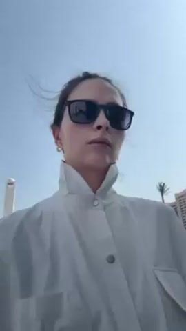 A woman with dark sunglasses and a white shirt is holding a camera looking upwards. The background shows tall buildings and a white-domed mosque. The sky is blue. The woman is walking.