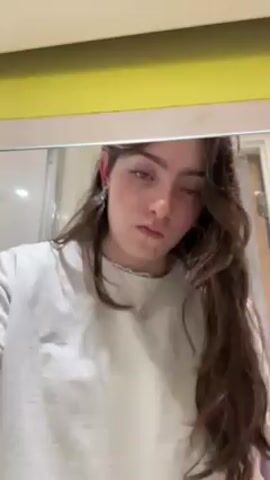 A young woman with long brown hair and light skin is shown from the chest up, facing a mirror. She is wearing a white sweatshirt and a necklace. She has a silver hoop earring in her left ear and painted fingernails. She sprays a bottle and then puts the sprayed liquid on a cotton pad. The background includes a light yellow surface above the mirror and a partially visible shower door to the left. The lighting is bright and even. The scene is still, except for the woman's movements.