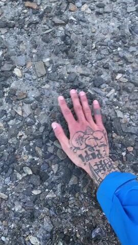 A person with tattoos on their left hand is breaking thin ice covering a body of water. The hand is fair-skinned, and the tattoos are black ink. The person is wearing a bright blue jacket. The water is a murky green-blue color, and the ice is thin and clear. The ground surrounding the water is rocky. The water is set against a mountain backdrop that has some snow. The sky is cloudy and gray.
