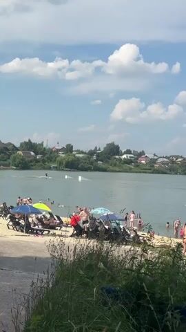 This video depicts a sunny day at a beach resort on a river. The water is calm and clear, with people swimming and engaging in water sports, such as wakeboarding. The beach itself is sandy, with several lounge chairs and umbrellas providing shade for sunbathers. Many people are present, enjoying the warm weather and recreational activities.

In the foreground, lush green grass and foliage create a natural border, hinting at a park or recreational area adjacent to the beach. The background reveals a picturesque scene of a small town nestled along the riverbank, with houses and trees adding to the tranquil atmosphere. The sky is a vibrant blue, dotted with fluffy white clouds, suggesting a pleasant summer day.

The overall mood of the video is one of leisure and enjoyment. People appear relaxed and happy, with families and individuals alike soaking up the sun and engaging in various activities. The presence of children's strollers suggests that this is a family-friendly destination.

The time of day appears to be midday or early afternoon, given the bright sunlight and the active nature of the crowd. The weather is clearly warm and sunny, ideal for a beach outing. The location is identified as Molokovo, Russia, a detail that adds specific geographic context to the otherwise universal scene of a summer beach day. There is no visible text in the video.