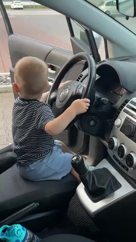 A young child is sitting in the driver's seat of a car, holding the steering wheel. The child is wearing a striped t-shirt and blue shorts. The car door is open. The child moves their hand to the ignition and pretends to turn the key. The child is looking at the steering wheel. There is a water bottle on the floor of the car.