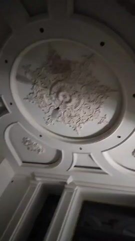 The video shows a low-angle view of a white ceiling with elaborate decorative moldings. The ceiling features a large, circular recessed section in the center, adorned with ornate plasterwork. Smaller, similarly decorated sections are visible in the surrounding areas. There are also several small, dark circular spots in the ceiling, possibly light fixtures. The video appears to be taken indoors.  The lower portion of the frame reveals elements of wall paneling and a framed object.