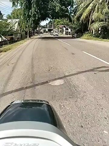 The video provides a first-person perspective from a scooter traveling along a paved road in Uleetanoh, Indonesia, during the daytime. The front part of the scooter's dashboard is visible in the foreground.

The road features some white markings and appears to be in a rural or semi-urban setting. On both sides of the road, various buildings, including houses and what appear to be small shops or stalls, are visible. Abundant green trees, including tall palm trees, line the road, casting shadows. Utility poles with electrical wires are also present.

Other vehicles, such as motorcycles and a white van, can be seen on the road. People are visible riding motorcycles and near the buildings. The sky is bright, indicating sunny weather. No discernible text is clearly visible in the video.