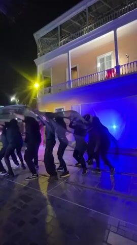 A group of approximately seven performers, dressed in black clothing and dark sneakers, are on an outdoor stage at night. They are holding a large, dark, flowing piece of fabric overhead, which billows and undulates as they move. Behind them is a two-story building with a balcony, illuminated by warm light. The stage is lit with vibrant blue and purple lights, creating a dramatic atmosphere. A bright yellow streetlight is visible to the left of the frame. The ground appears to be paved with dark stones or bricks.
